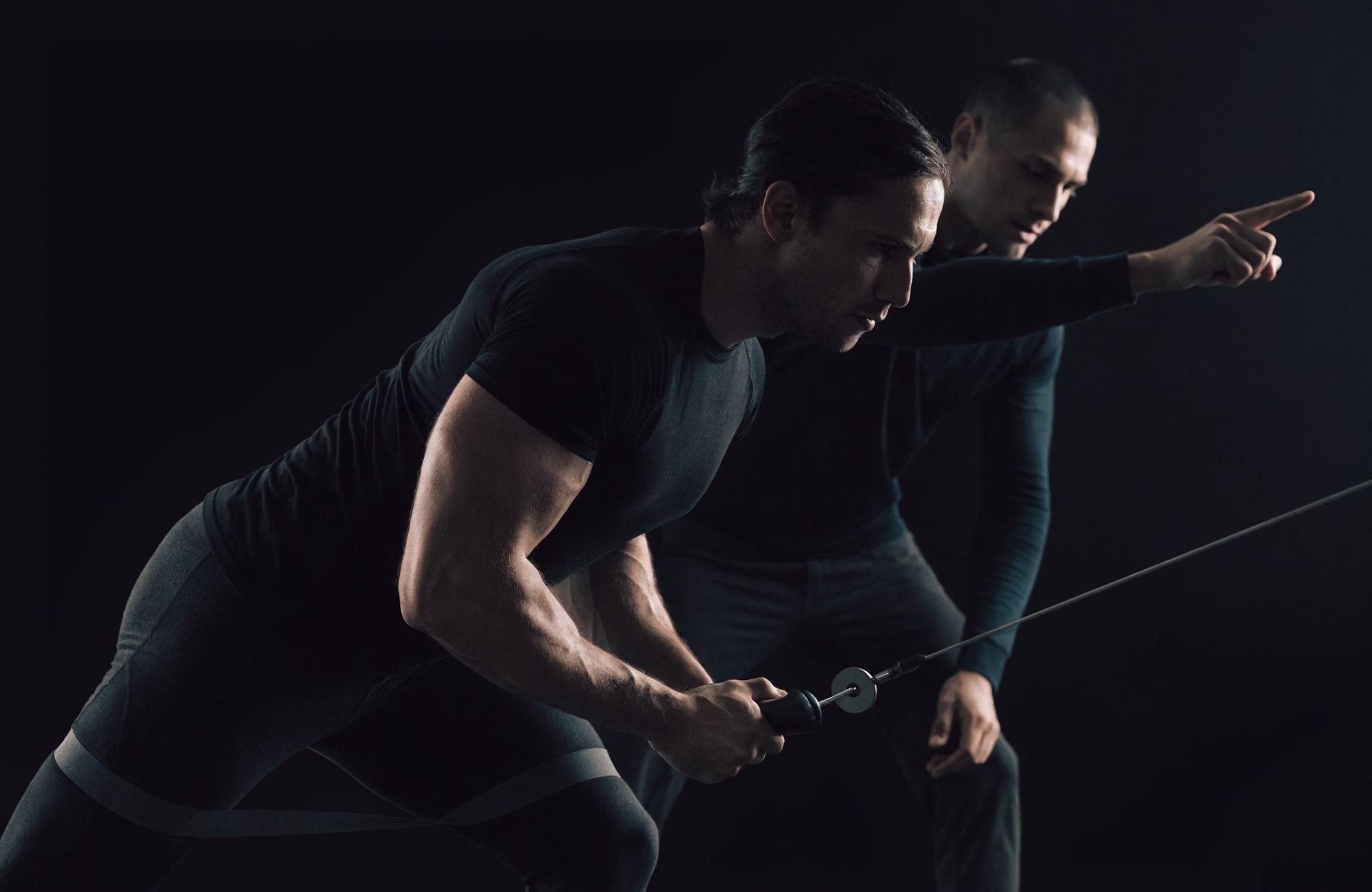 A male client grips a cable handle and drives through a resisted lateral movement while a trainer directs him in a dark performance gym, the kind of footwork that carries onto the pickleball court.